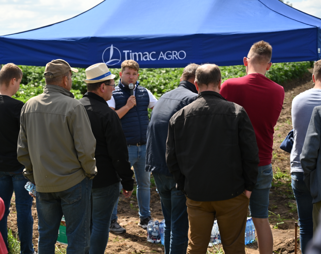 Grupa rolników słucha mężczyzny trzymającego mikrofon pod niebieskim namiotem z logo "Timac Agro". Namiot jest ustawiony na polu ziemniaków, a uczestnicy są uważnie skupieni na prezentacji.