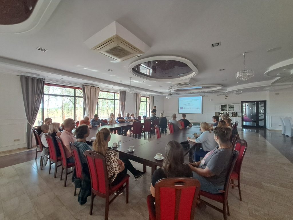 Grupa uczestników wyjazdu studyjnego zasiadająca przy dużym stole konferencyjnym w eleganckiej sali. Na ekranie widoczna jest prezentacja dotycząca projektu współfinansowanego przez Unię Europejską. Uczestnicy uważnie słuchają prelegenta, atmosfera jest skupiona, sprzyjająca wymianie wiedzy.