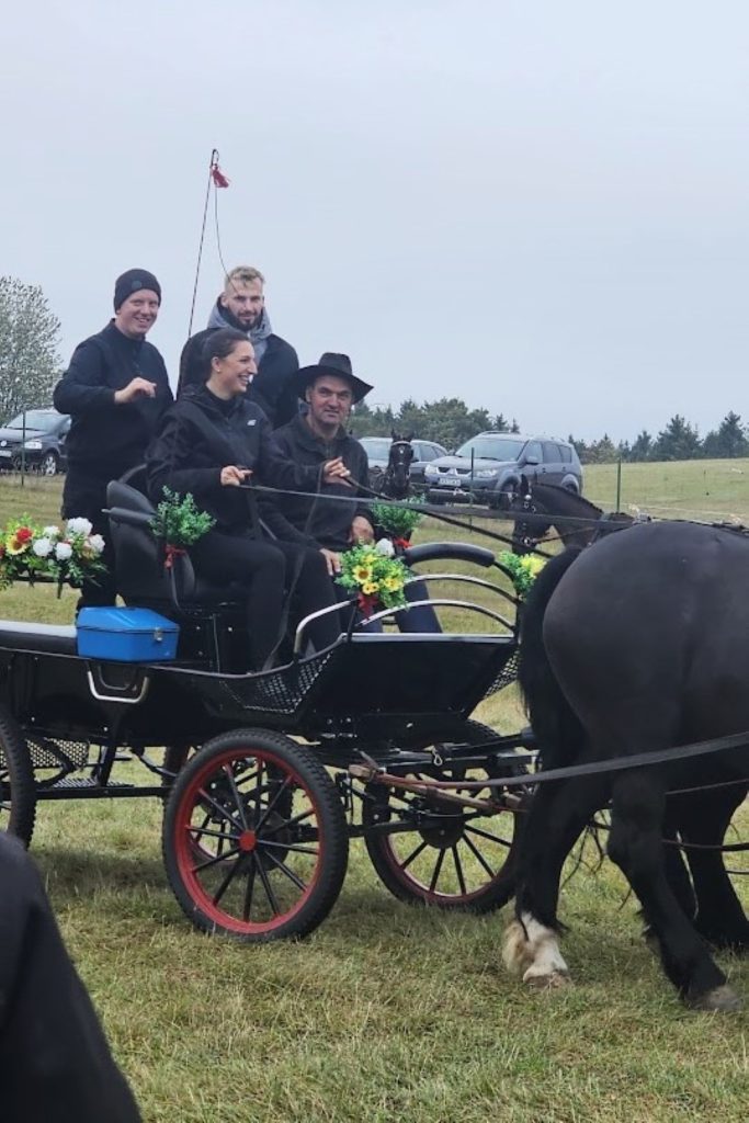 Zaprzęg dwukonny z powożącą i luzakami w bryczce przystrojonej kwiatami.