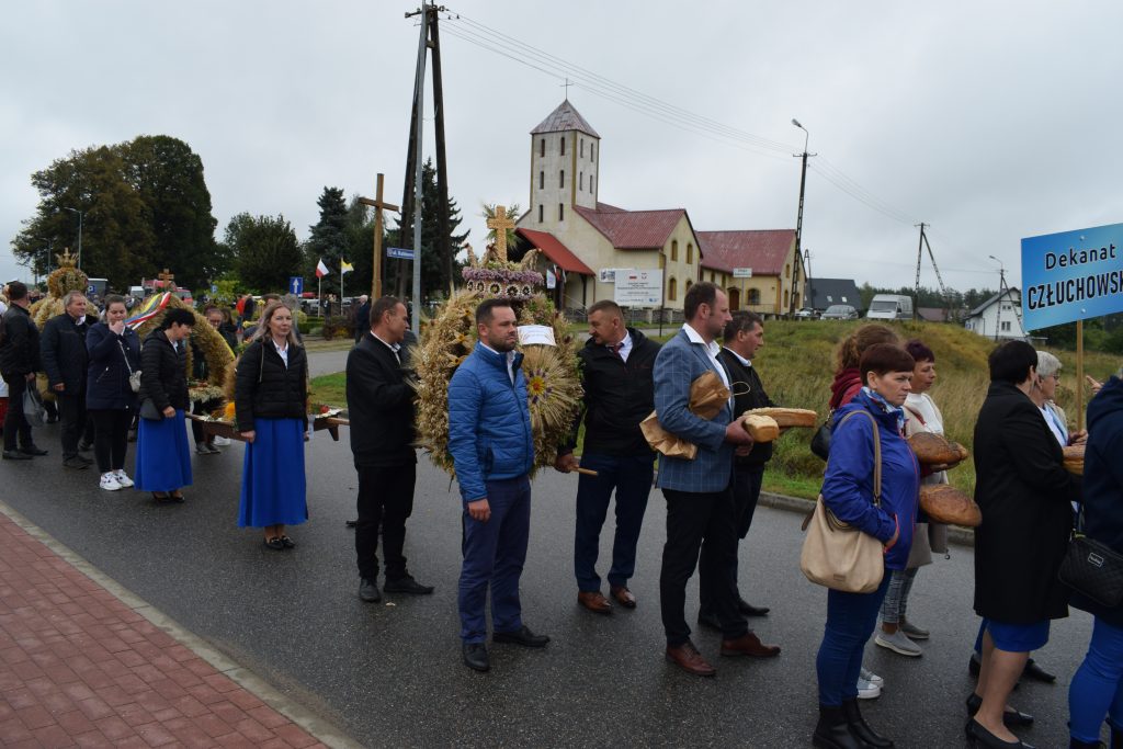 Uczestnicy niosą wieńce dożynkowe, symbolizujące dziękczynienie za plony, a w tle widać lokalnych mieszkańców, którzy podziwiają to tradycyjne wydarzenie.