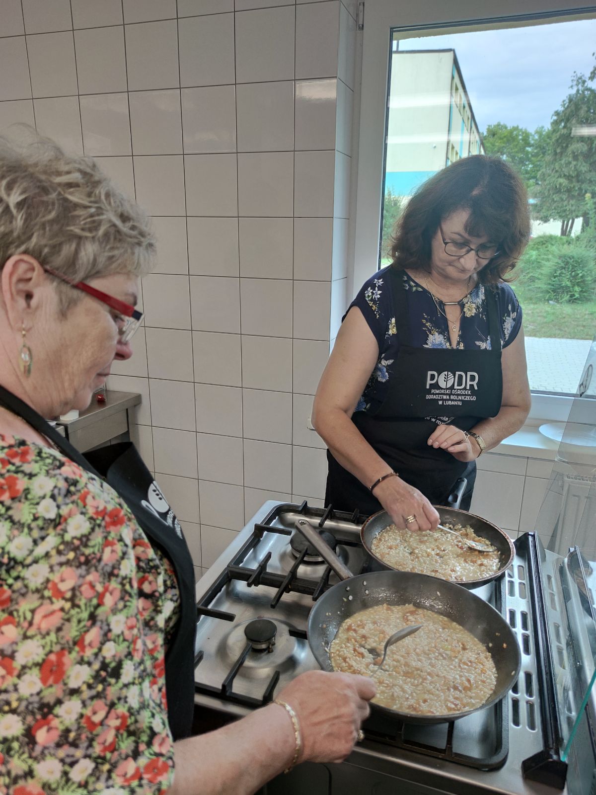 Dwie kobiety biorące udział w warsztatach kulinarnych w trakcie przygotowywania potraw na kuchence. Obie mają na sobie fartuchy z logo Pomorskiego Ośrodka Doradztwa Rolniczego (PODR). W garnkach widać risotto, które jest starannie mieszane. W tle widoczny jest krajobraz za oknem oraz wnętrze nowoczesnej kuchni warsztatowej.