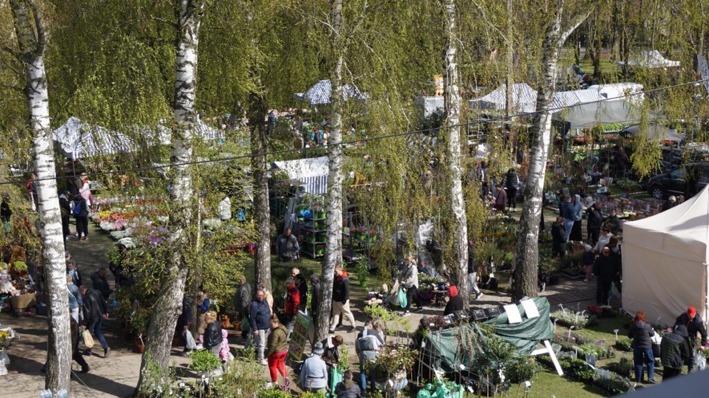 Widok z góry na teren targów ogrodniczych; liczne stoiska i tłumy odwiedzających wśród drzew.