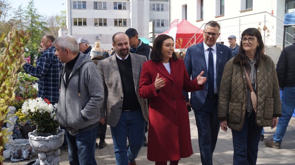 Grupa uczestników, w tym kobieta w czerwonym płaszczu, spaceruje alejką targową; rozmowy i zwiedzanie stoisk.