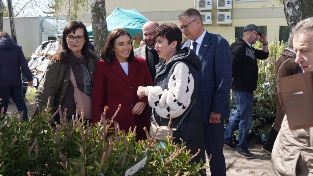 Grupa gości, w tym kobieta w czerwonym płaszczu, ogląda rośliny ozdobne na stoisku ogrodniczym podczas targów.