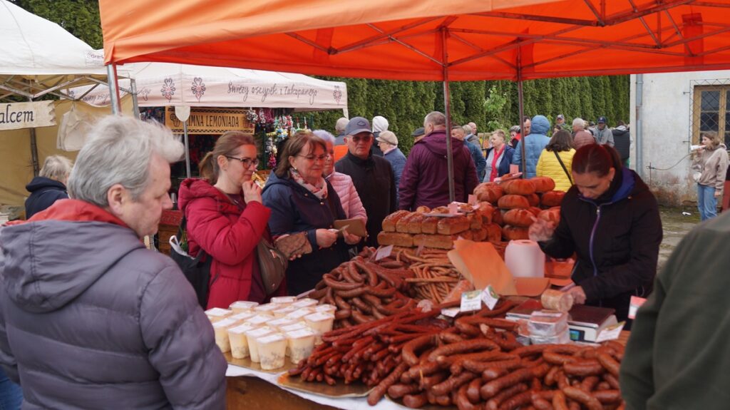Odwiedzający kupują wędliny na stoisku pod czerwonym namiotem; sprzedawcy obsługują klientów.