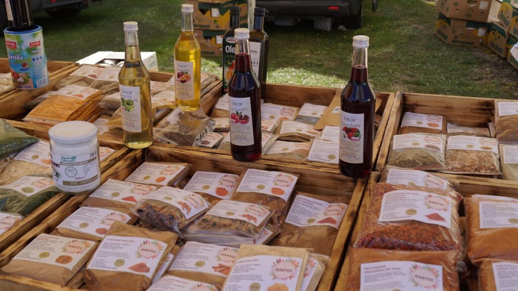 Stoisko z przyprawami w woreczkach oraz butelkami z produktami regionalnymi, ustawione na drewnianym stole.