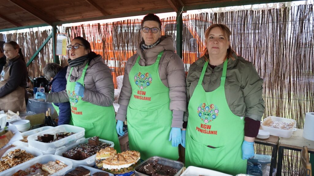 Stoisko Koła Gospodyń Wiejskich; kilka osób stoi za stołem z domowymi produktami i przetworami.