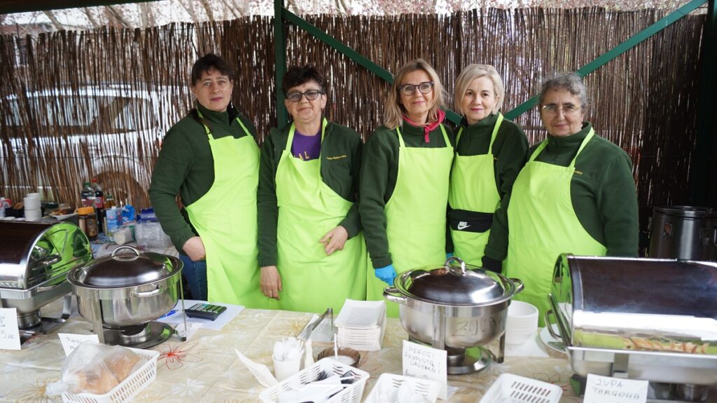 Stoisko Koła Gospodyń Wiejskich; kilka osób stoi za stołem z domowymi produktami i przetworami.