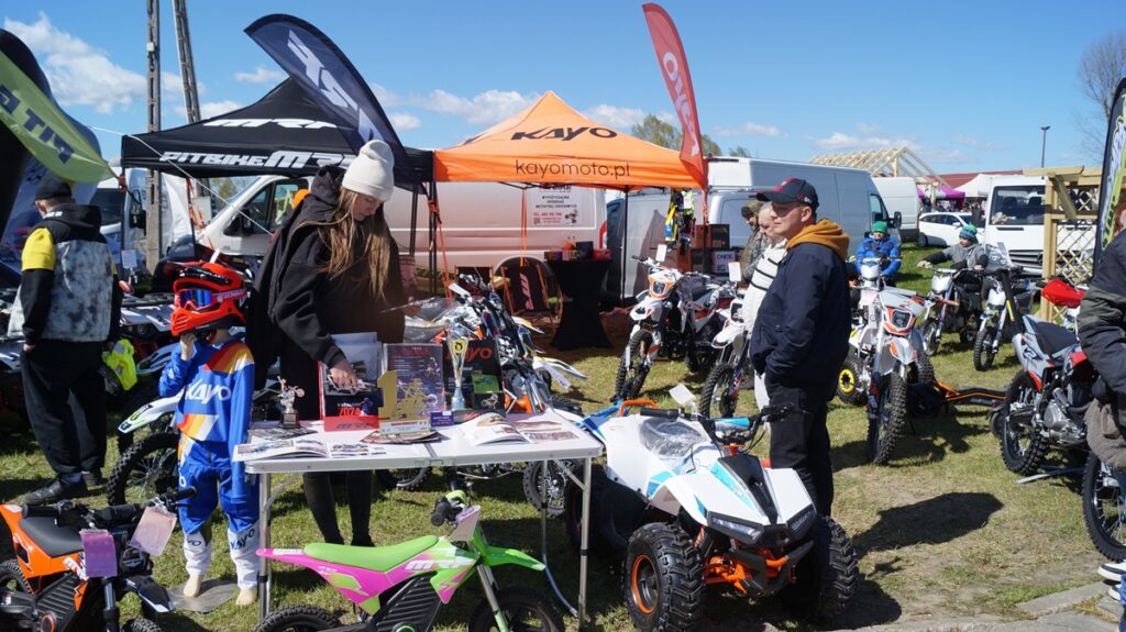 Stoisko z pojazdami rekreacyjnymi i sprzętem – motocykle, quady oraz akcesoria prezentowane na świeżym powietrzu.