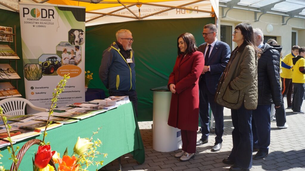 Stoisko promocyjne z materiałami informacyjnymi; kilka osób rozmawia z wystawcą przy ladzie przykrytej zielonym obrusem.