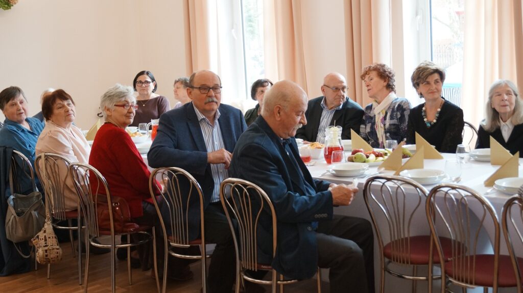 Emerytowani pracownicy ośrodka siedzą przy stołach podczas spotkania w sali.