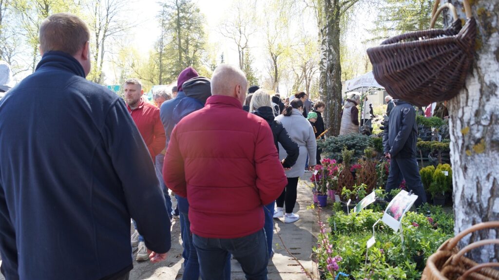 Odwiedzający spacerują alejką targową między stoiskami z roślinami; w tle drzewa i namioty wystawców.