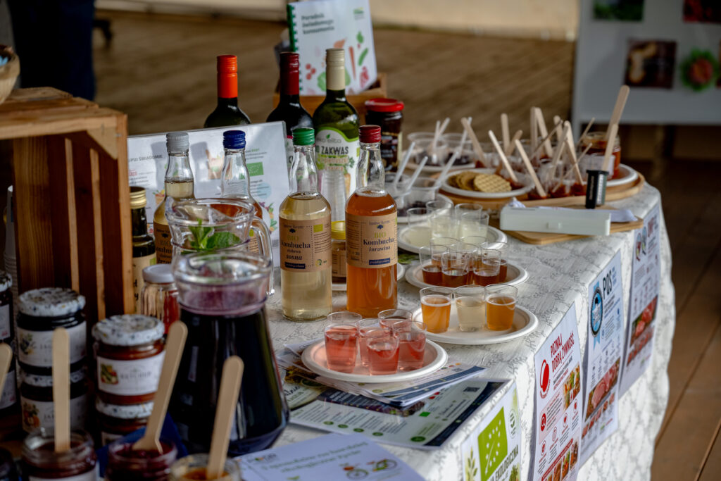 Zbliżenie na stół degustacyjny w strefie edukacyjnej #KupujŚwiadomie z certyfikowanymi produktami lokalnych producentów, napojami, przetworami oraz materiałami informacyjnymi dotyczącymi systemów jakości żywności.