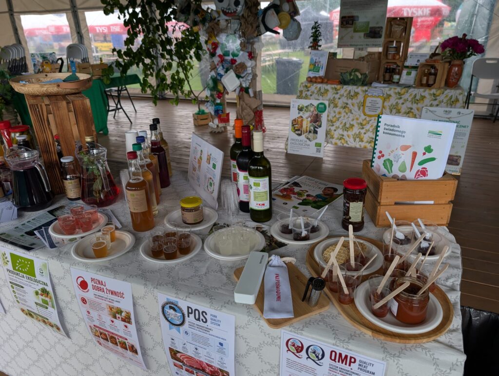 Stół degustacyjny w strefie #KupujŚwiadomie z produktami lokalnymi, przetworami, napojami i materiałami edukacyjnymi dotyczącymi systemów jakości żywności.